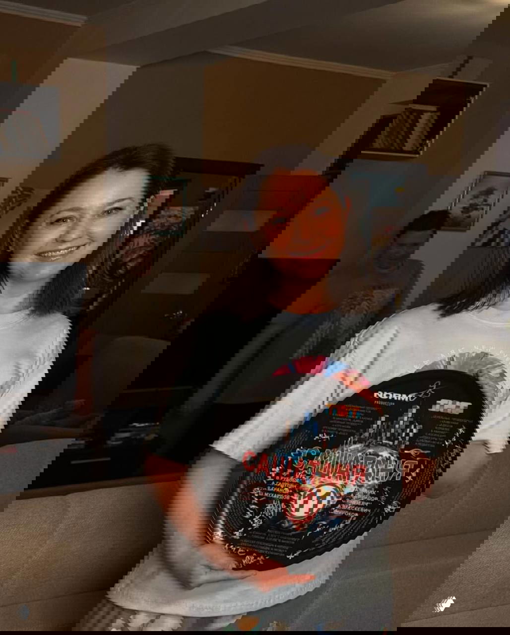 Валентина Рубцова на фоне гостиной держит диск с символикой «САШАТАНЯ 11», позади видны другие участники съёмок.