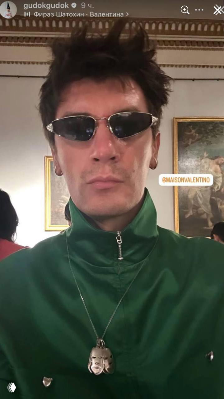 Селфи мужчины в зелёной рубашке с крупным кулоном на шее и темными очками, сделанное внутри зала показа мод рядом с картинами на стене.