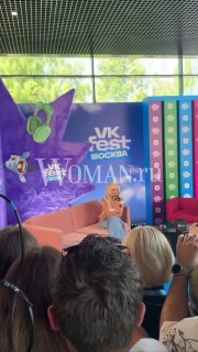 Ида Галич сидит на сцене фестиваля на фоне брендированной панели VK Fest и Woman.ru; в кадре видны часть публики и камеры, атмосферный ракурс.
