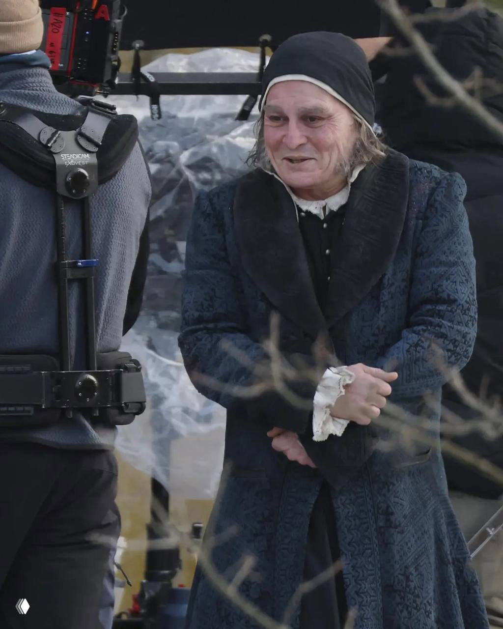 Актёр в гриме XIX‑векового персонажа на улице: сценический костюм с кружевными манжетами, окружающая съёмочная техника и члены команды.