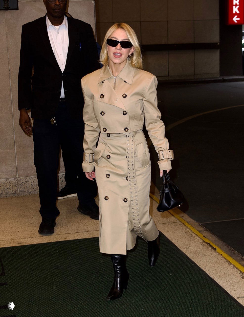 Женщина в двубортном бежевом тренче с поясом и декоративными клёпками, чёрная сумка и сапоги — стильный городской образ.