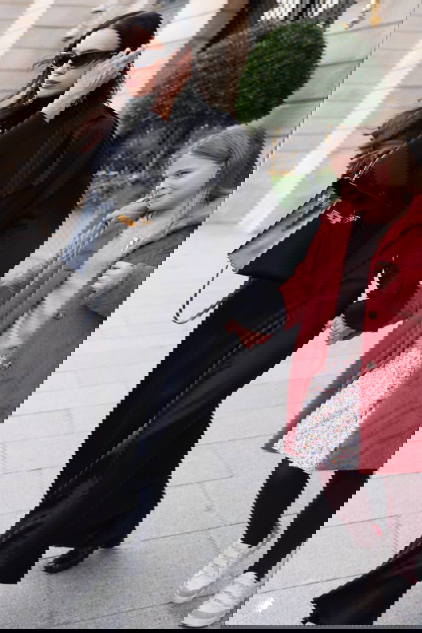 Женщина в чёрном платье держит за руку девочку в розовом пальто и цветочном платье, они идут по парижской улице; повседневный папарацци-кадр.