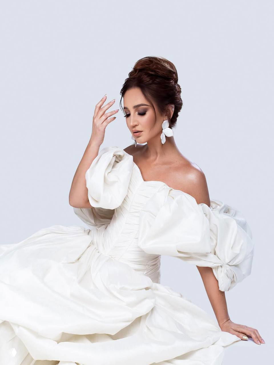 Женщина в белом корсетном платье в сидящей позе, взгляд вниз, акцент на макияже, украшениях и сложной фактуре юбки на нейтральном фоне.