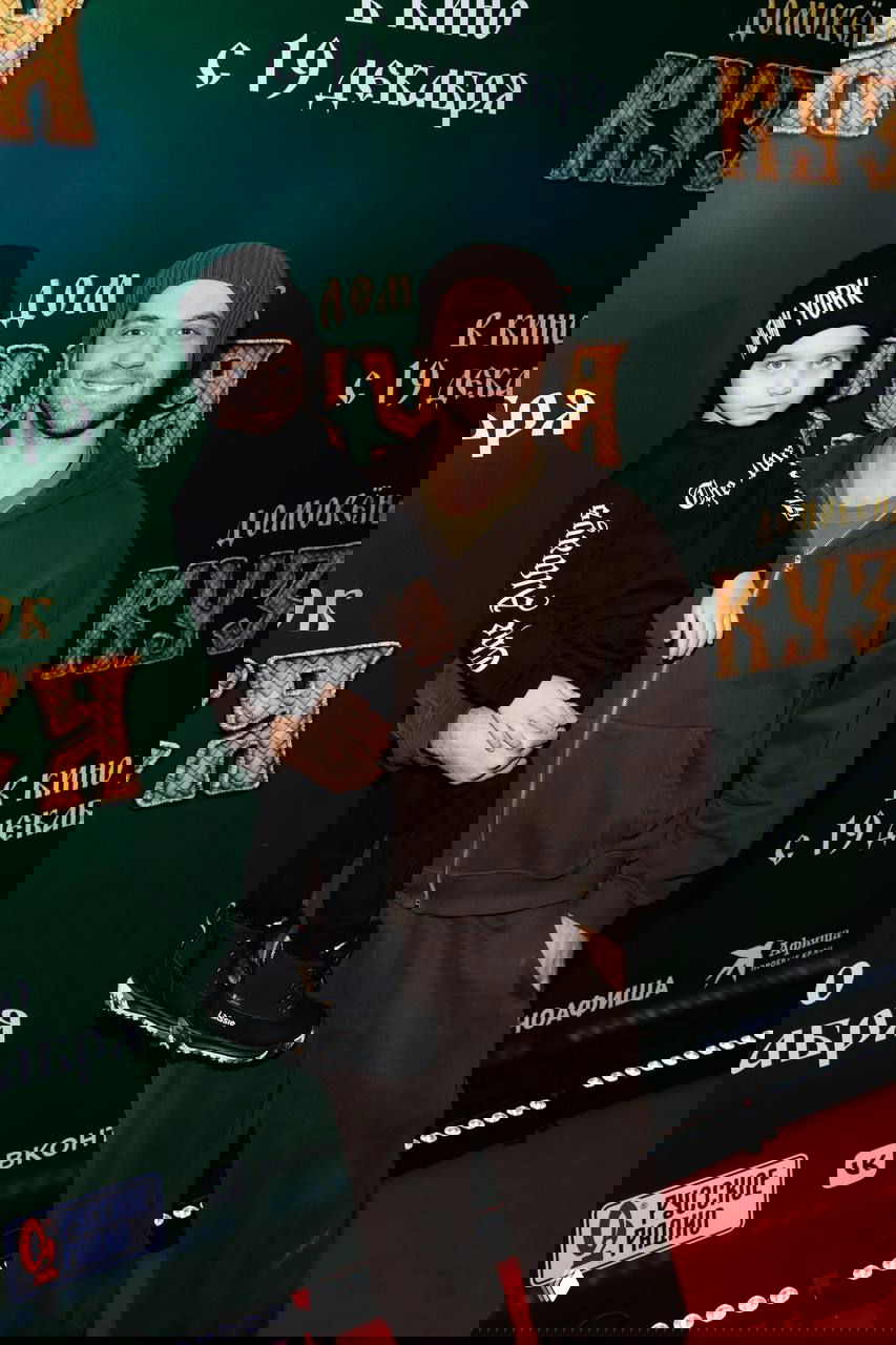 Мужчина в тёплой одежде и шапке держит ребёнка на фоне постера фильма; кадр с премьерной фотозоны, дружелюбная сцена на красной дорожке.