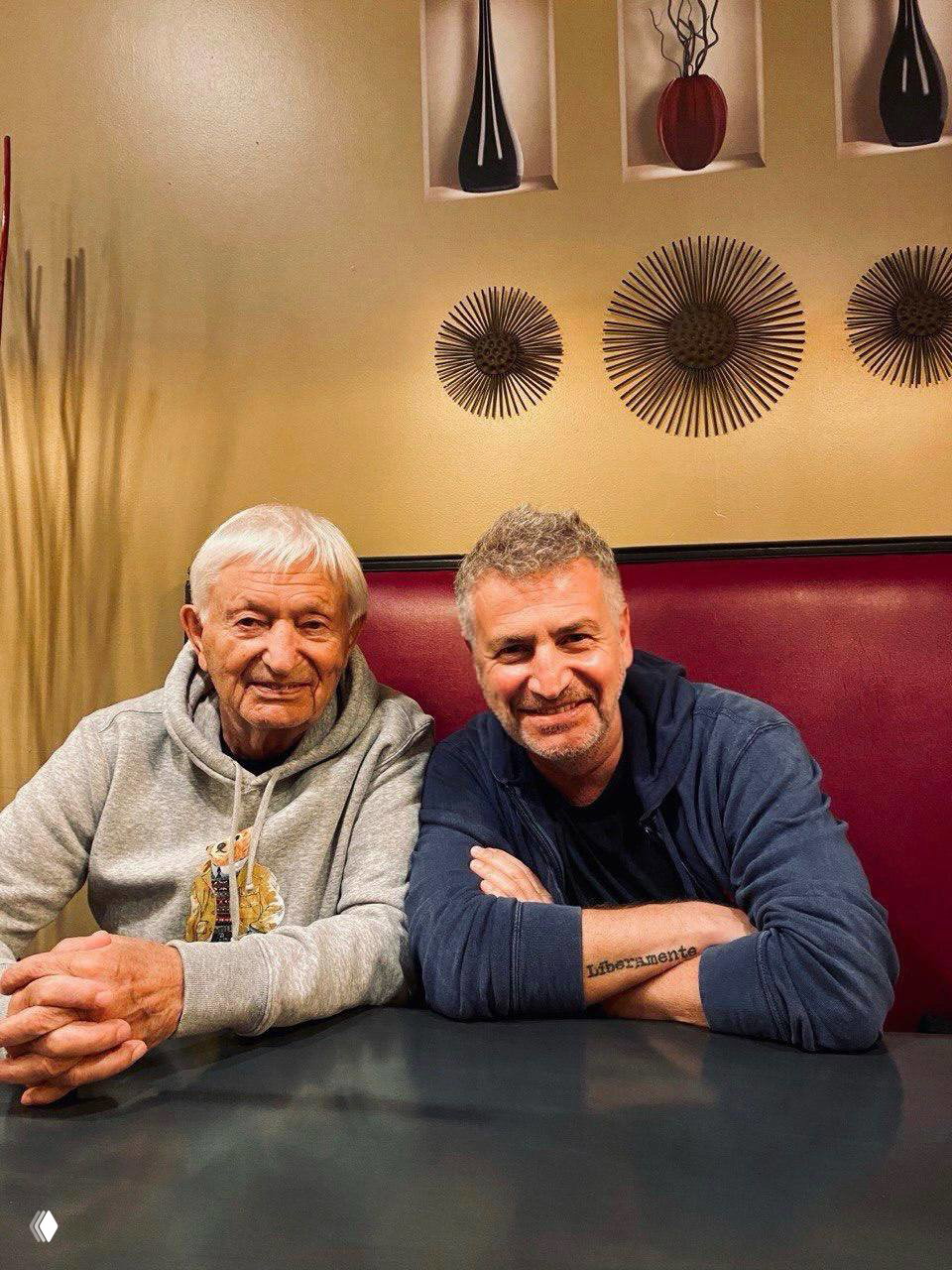 Два мужчины за столом в кафе: пожилой мужчина и мужчина среднего возраста, тёплая непринуждённая атмосфера и декор на стене.