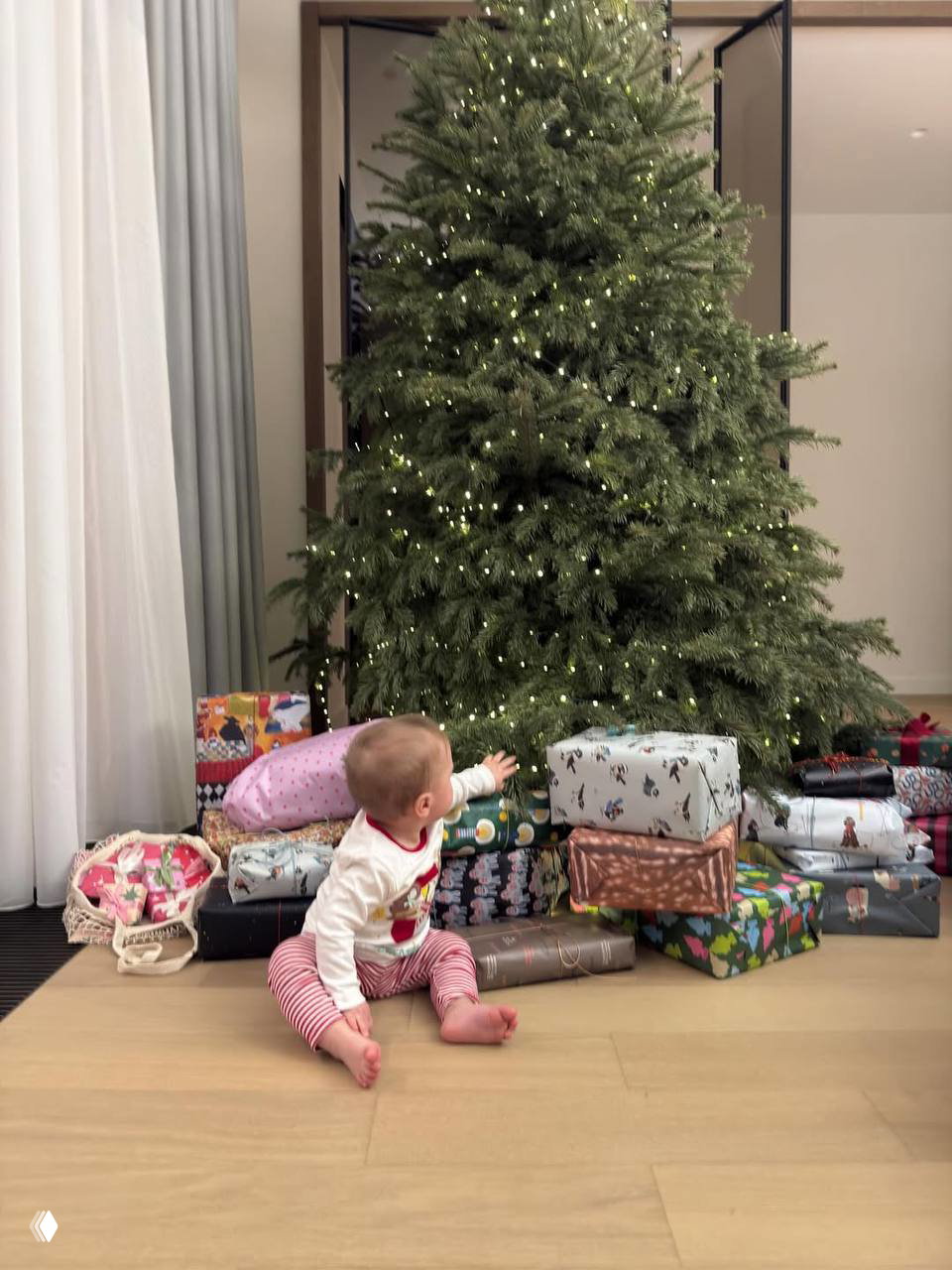 Малыш на полу у ёлки тянется к подаркам: много упакованных коробок у большой украшенной рождественской ели в домашнем интерьере.