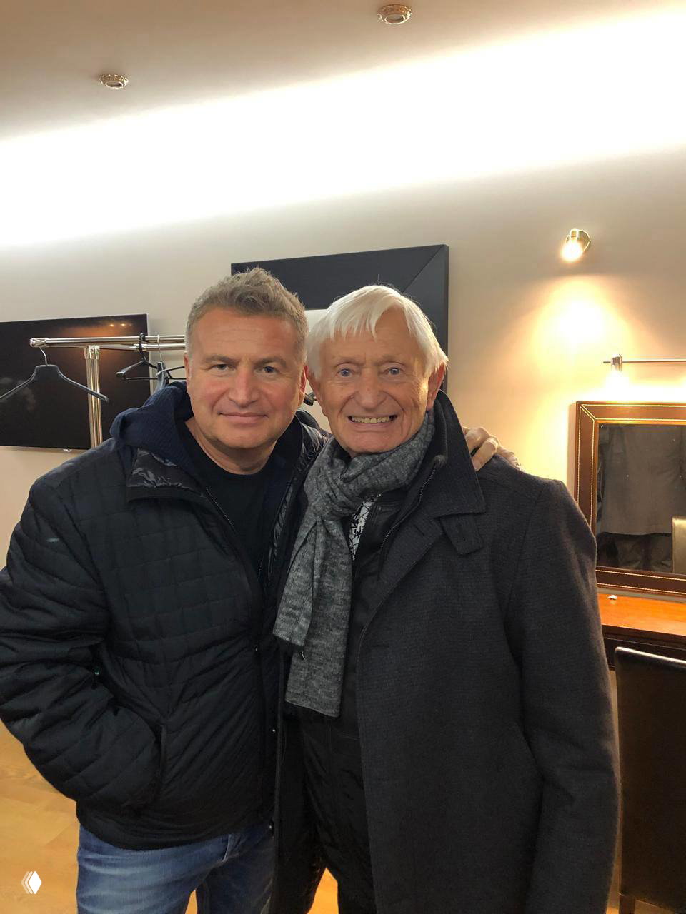 Два мужчины в тёплой одежде в гримёрке: пожилой мужчина и спутник среднего возраста, они стоят рядом и улыбаются в камеру.