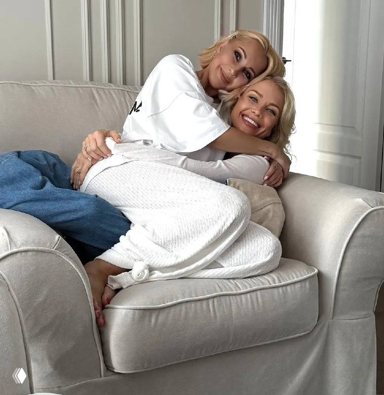 Две улыбающиеся женщины в светлом интерьере сидят на уютном диване, обнимаются и выглядят близкими и расслабленными в домашней обстановке.