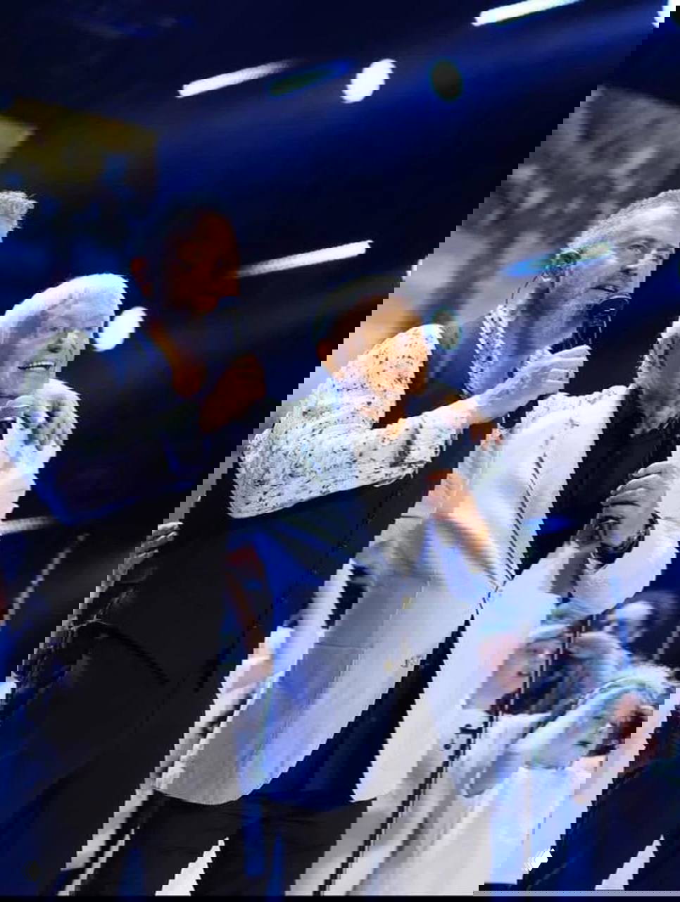 Два исполнителя на сцене с микрофонами: пожилой мужчина в светлом пиджаке и партнёр, яркое сценическое освещение и дети на заднем плане.