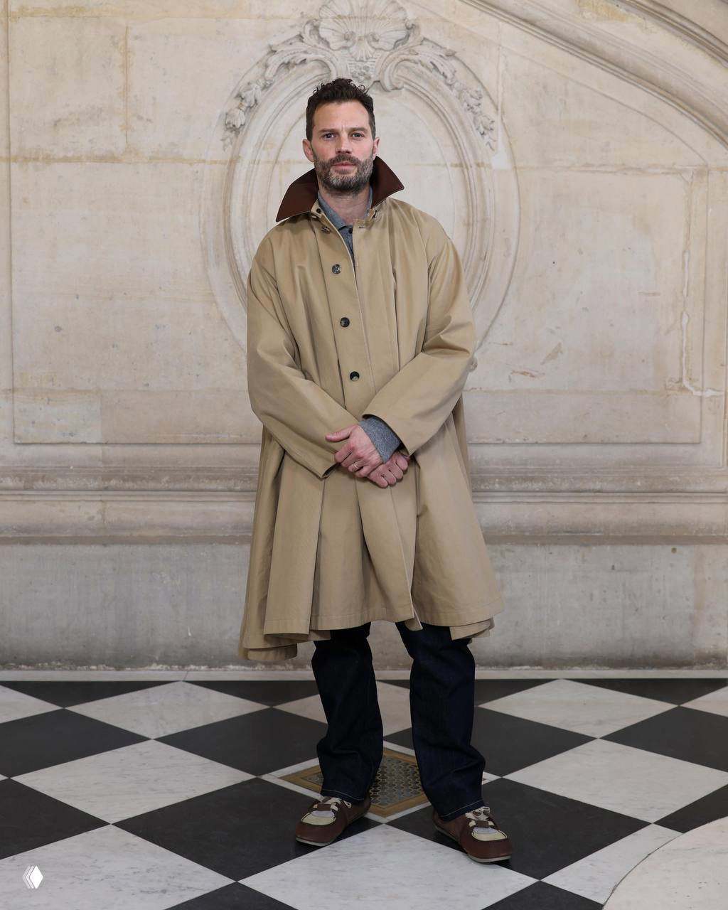 Мужчина в длинном бежевом тренче с поднятым воротником, джинсах и кедах на мраморном фоне — классический верхний слой в нейтральных тонах.