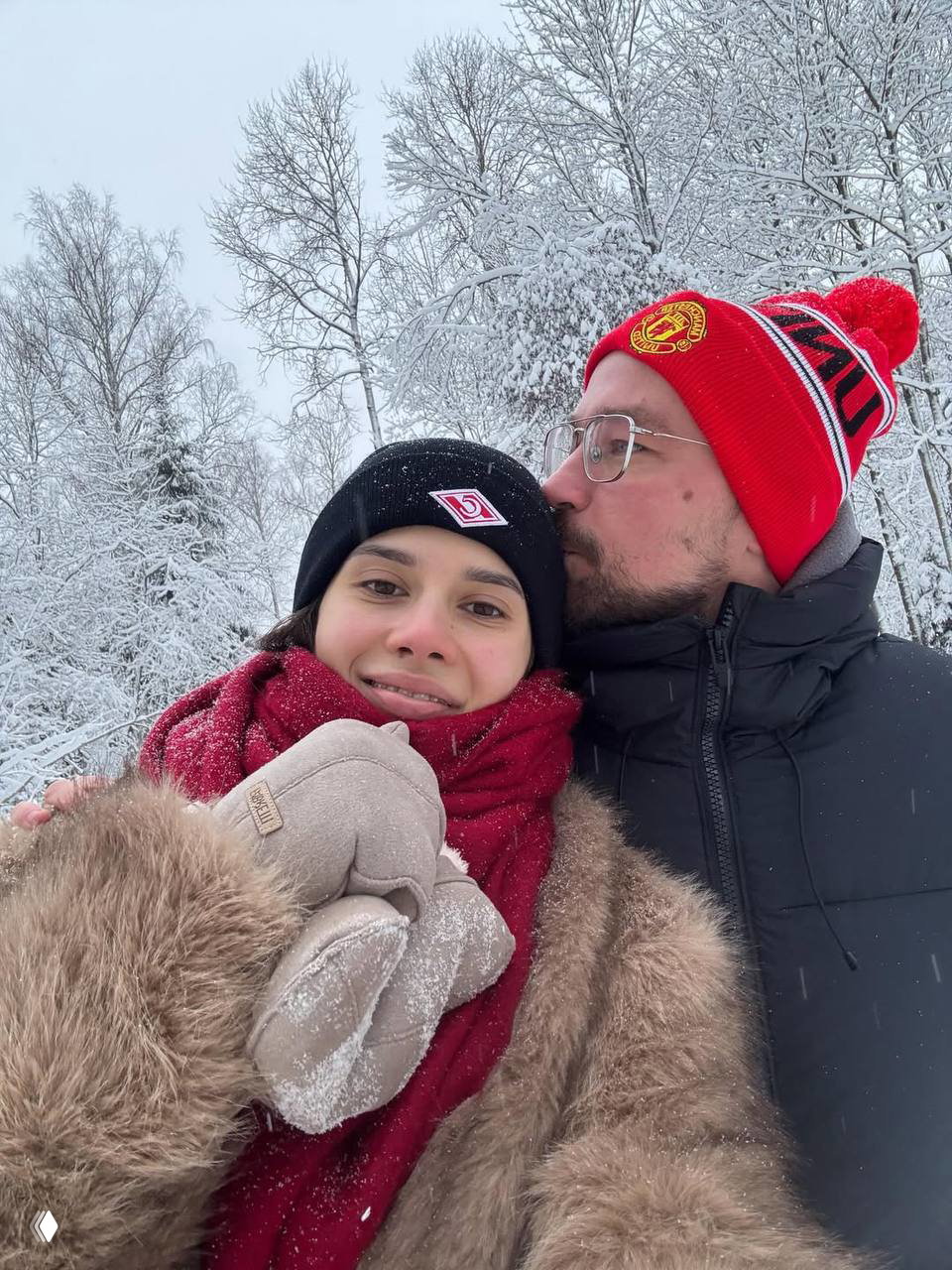Селфи пары на прогулке: Виктория и Александр в тёплой зимней одежде на фоне заснеженного леса, мужчина целует жену в щёку.