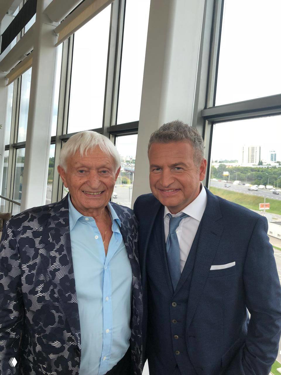 Двое мужчин в деловых костюмах у большого панорамного окна: пожилой мужчина и мужчина среднего возраста позируют в светлом зале.