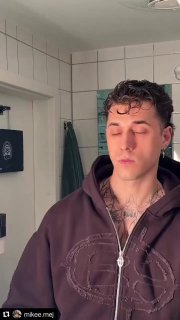 Крупный план молодого человека в худи в санузле с плиткой на стене, глаза полузакрыты, видна цепь и тату на шее, бытовой фон.