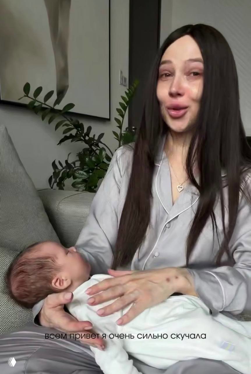 Валерия в длинном тёмном парике с заплаканным лицом держит младенца на руках, сидя на диване в домашней атмосфере; лицо выражает эмоции после лечения.