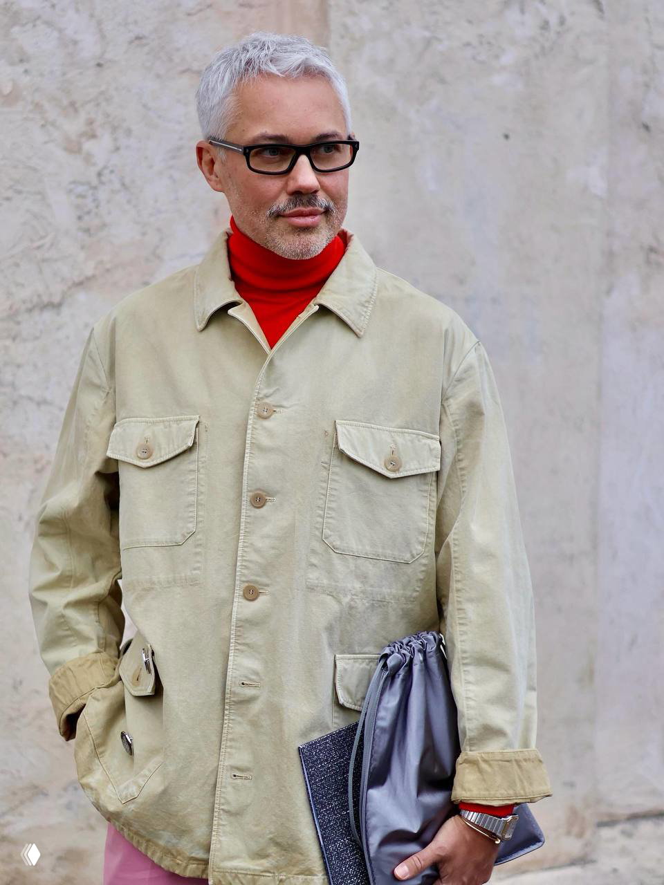 Мужчина в бежевой рабочей куртке и яркой красной водолазке, с папкой в руках, на нейтральном фоне — современный аккуратный образ.