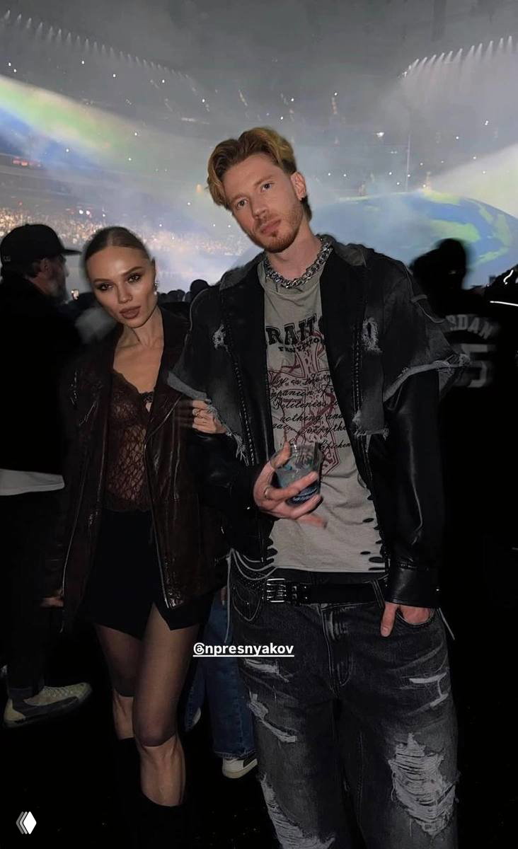 Пара на концерте: мужчина и женщина в модных нарядах стоят в толпе на арене с разноцветной сценической подсветкой и большой аудиторией на заднем плане.