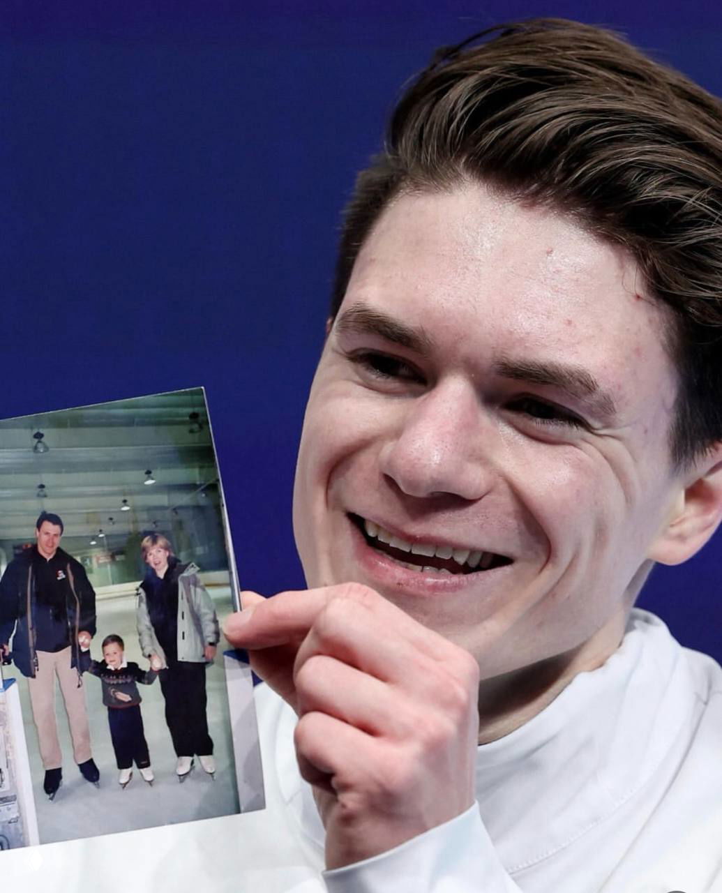 Крупный план фигуриста Максима Наумова, улыбающегося и держащего в руках семейную фотографию родителей на фоне олимпийской арены, снимок семьи в руках.