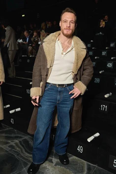 Мужчина в коричневом дубленом пальто с меховой отделкой и джинсах сидит в ряду показа — тёплый многослойный образ в сезонном ключе.