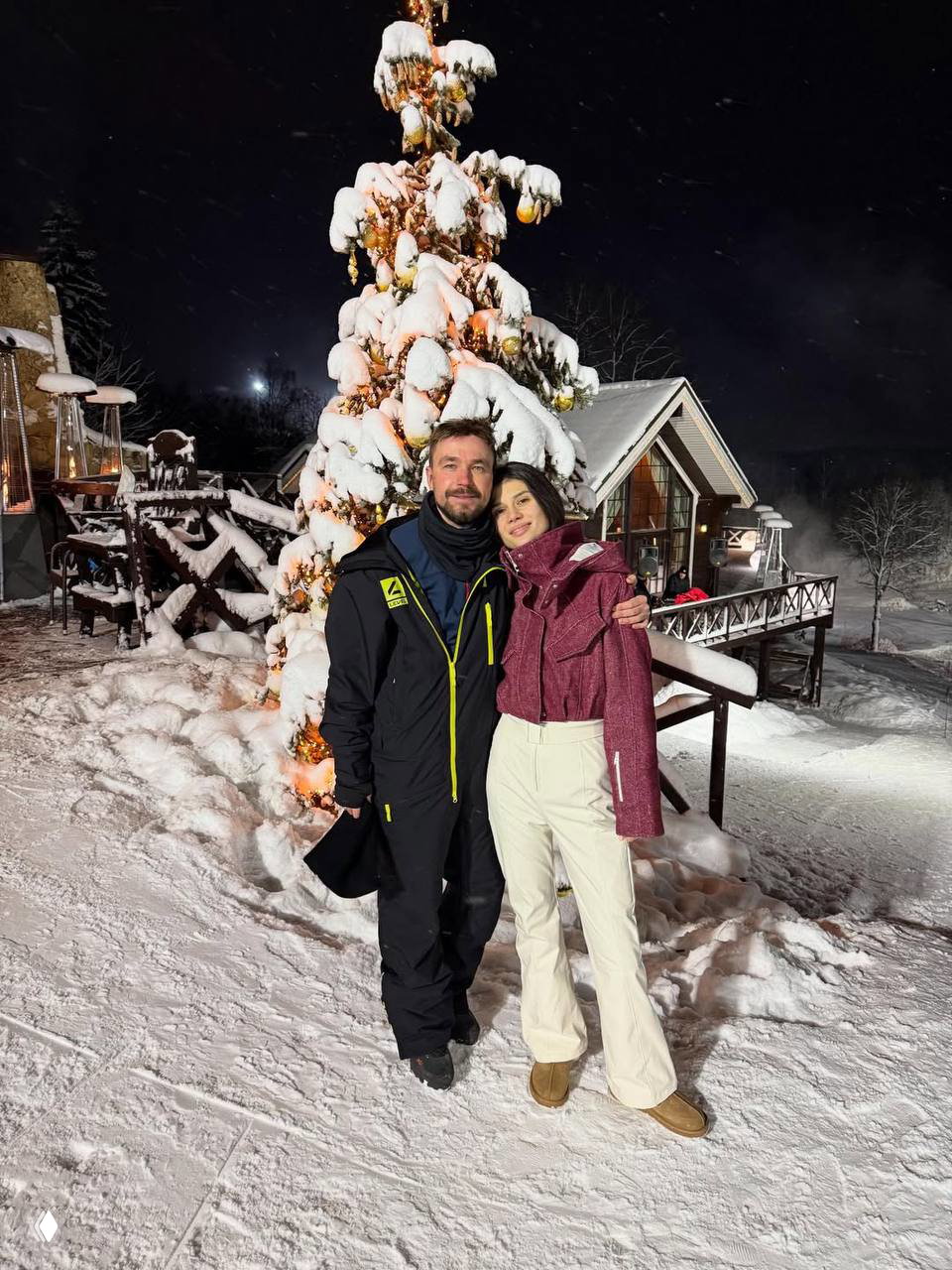 Пара обнимается у украшенной снежной ёлки ночью возле загородного дома: вокруг заснеженные деревья и тёплое вечернее освещение.