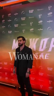 Мужчина в тёмной рубашке и пиджаке позирует на фоне баннера с брендингом фильма, сцена украшена цветной подсветкой.