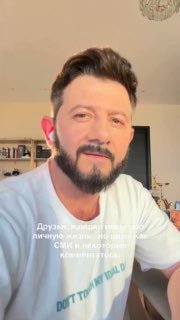 Крупный план Михаила Галустяна в домашнем видео: светлая футболка, наушник и книги на полке в интерьере за ним.