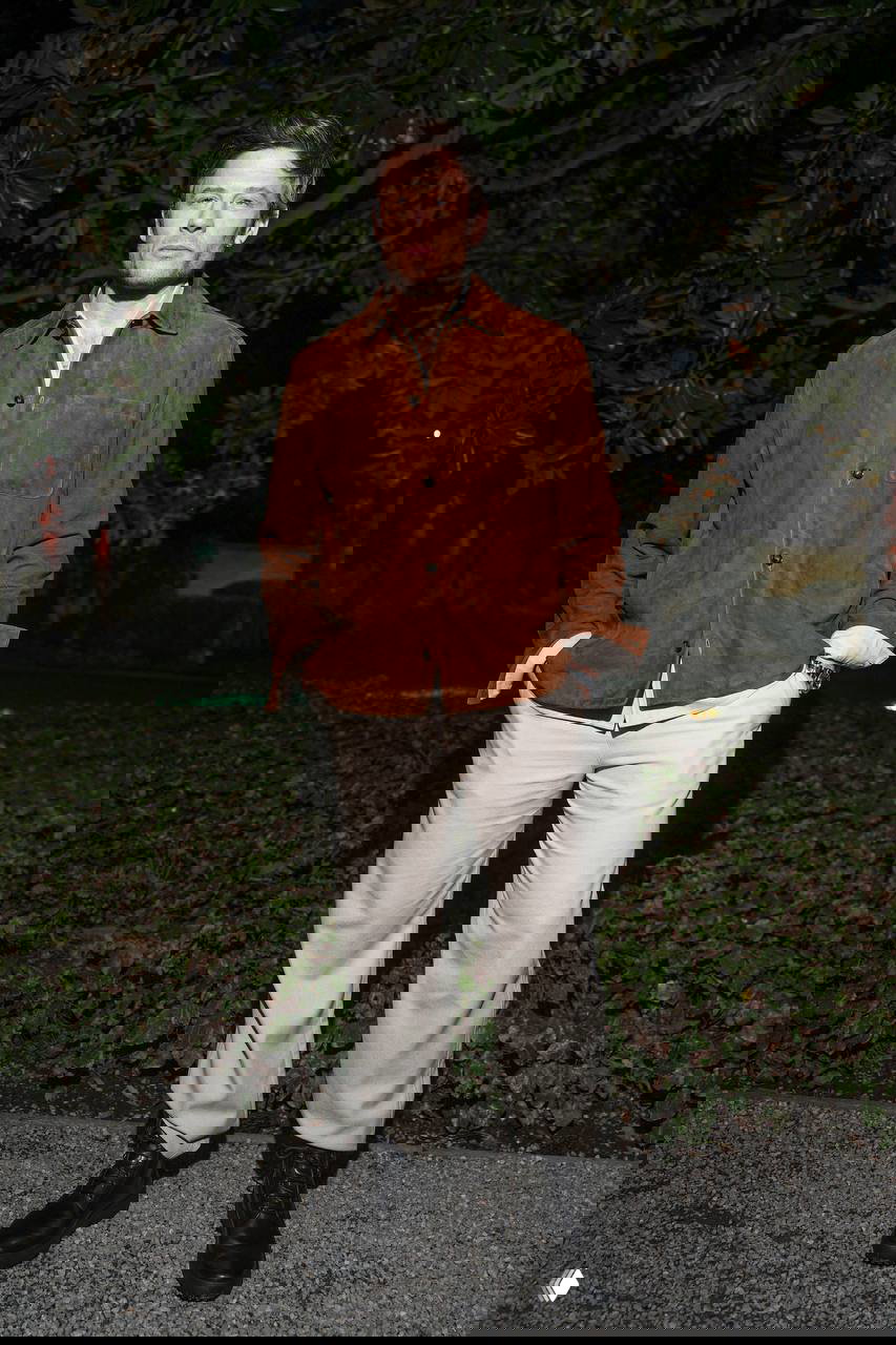 Мужчина в коричневой замшевой рубашке и светлых брюках стоит в парковой зоне вечером — неформальный вечерний образ с грубой обувью.