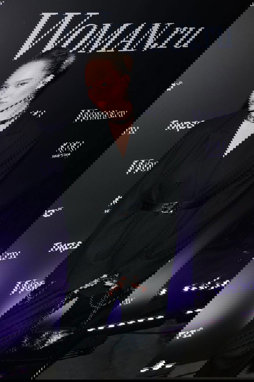 Гостья в черном костюме с поясом и массивными украшениями позирует у брендволла Woman.ru во время вечеринки.