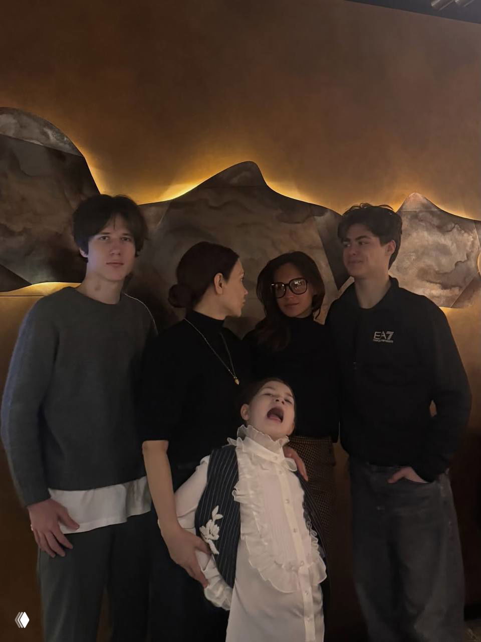 Фотография в тёплом интерьерном свете: взрослая женщина, подростки и маленькая девочка в нарядной блузке, одна поза исполнена шутливости