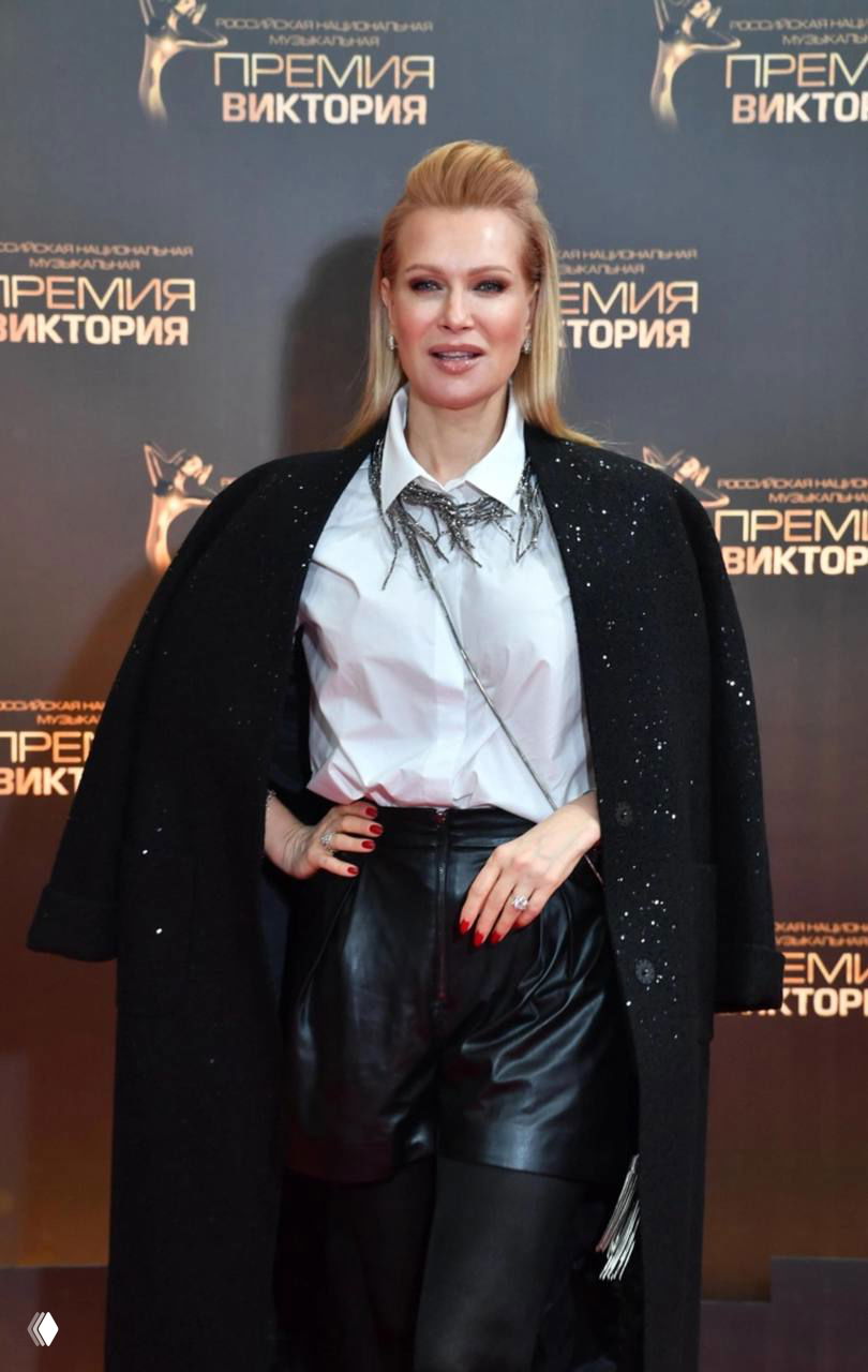 Светловолосая актриса в белой блузке и чёрном пальто позирует у медийной стены на церемонии, полноростовой портрет в интерьере мероприятия.