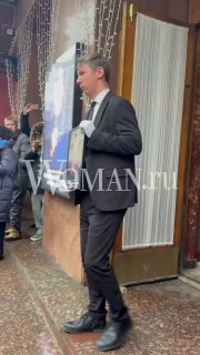 Мужчина в тёмном костюме несёт портрет покойного актёра во время церемонии прощания у входа; рядом люди в трауре и декоративные гирлянды на стене.
