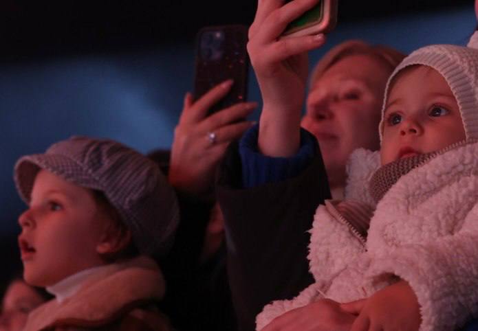 Два ребёнка в тёплой одежде на руках у взрослых при свете сцены внимательно следят за представлением; на заднем плане зрители с телефонами.