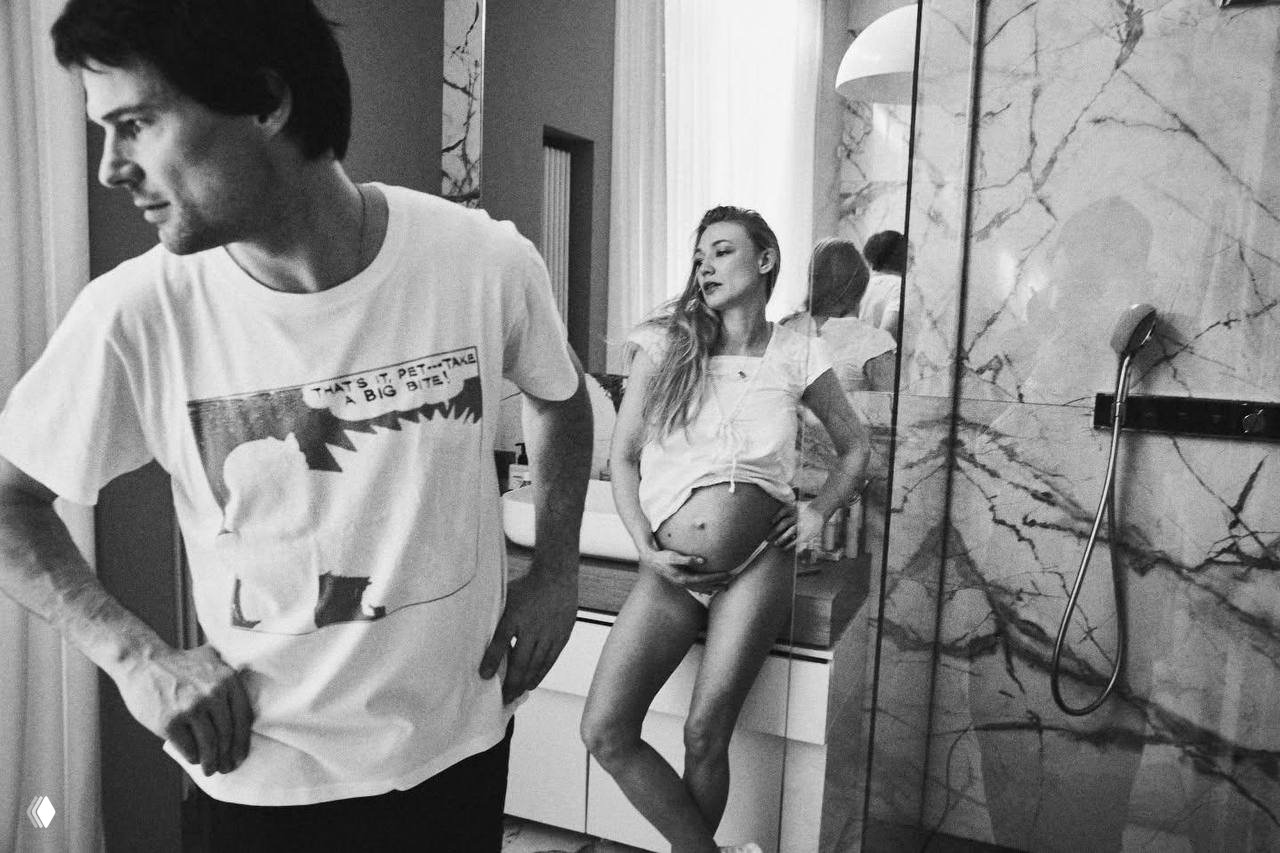 Чёрно‑белая фотография в интерьере дома: мужчина в футболке на переднем плане, женщина с округлившимся животом стоит у раковины в ванной комнате.