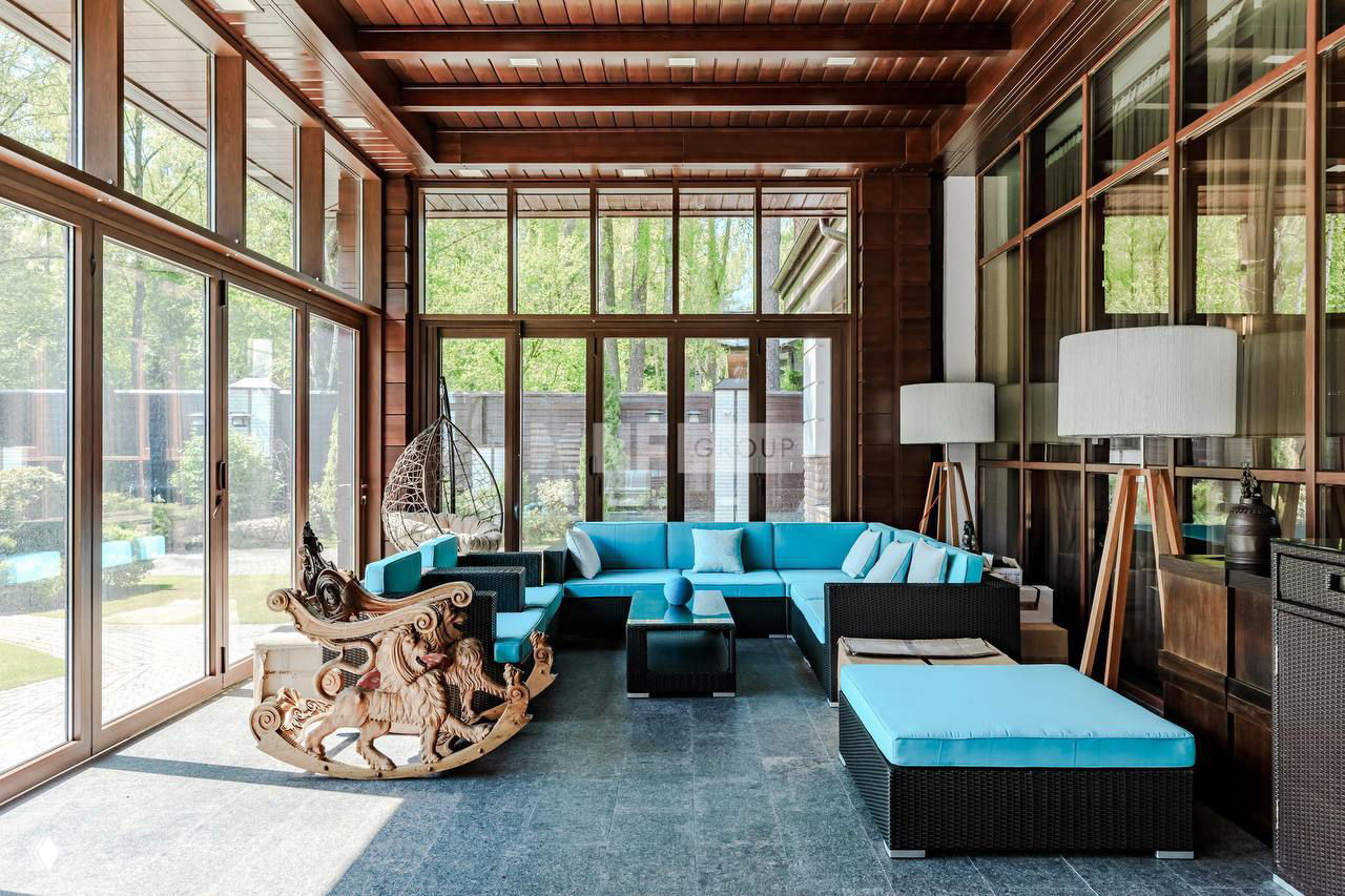 Светлая веранда с панорамными окнами и бирюзовой плетёной мебелью на фоне деревянной отделки — уютная зона отдыха в хозяйском доме особняка.