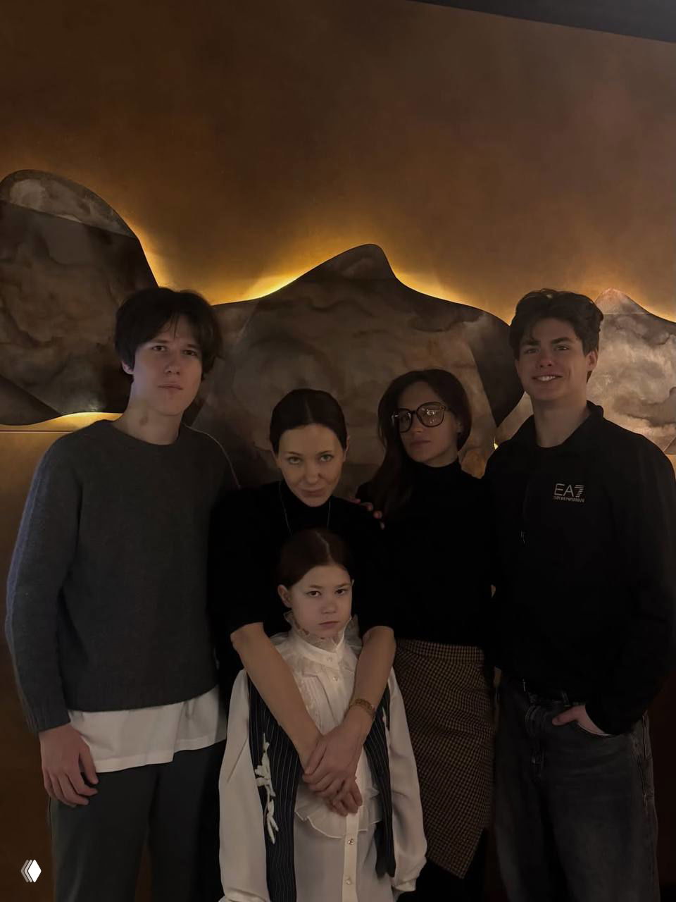 Портретная группа: несколько подростков и женщина стоят вместе в тёплом интерьере, младшая девочка посередине выглядит серьёзной