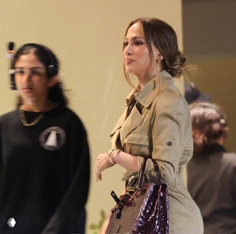 Крупный план Джей Ло в профиль: аккуратная низкая причёска, золотые украшения и часы, она держит лакированную сумку перед спектаклем.