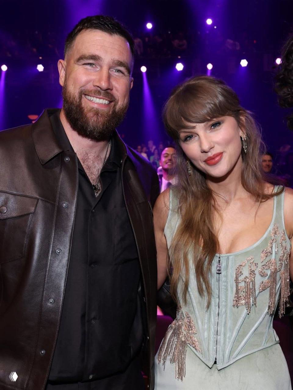 Две персоны в зале церемонии: женщина в платье и мужчина в тёмной куртке улыбаются на фоне сценического освещения и зрителей.