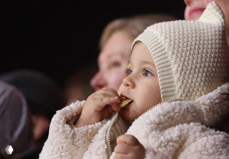 Крупный план малыша в уютном вязаном комбинезоне и шапочке, лицо выражает восхищение и удивление от происходящего на льду.