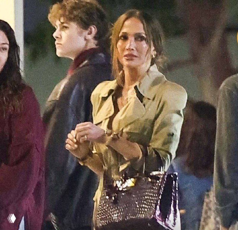 Джей Ло в толпе зрителей смотрит в камеру; видно верхнюю часть её образа, складки тренча и блестящую крокодиловую сумку в руках.