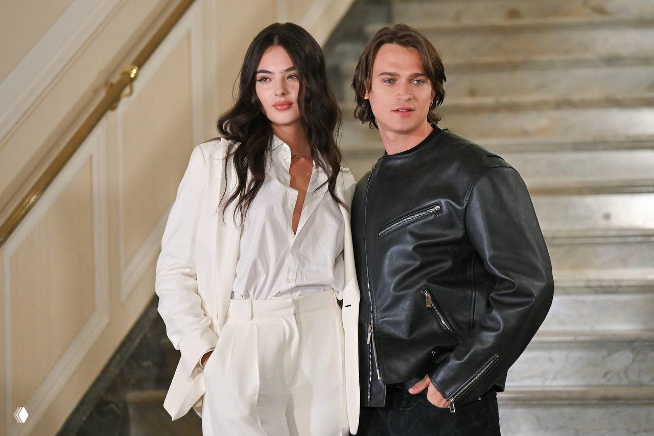 Дева Кассель в белом костюме и Сол Нанни в кожаной куртке на светском мероприятии: лестничный фон и официальная атмосфера.