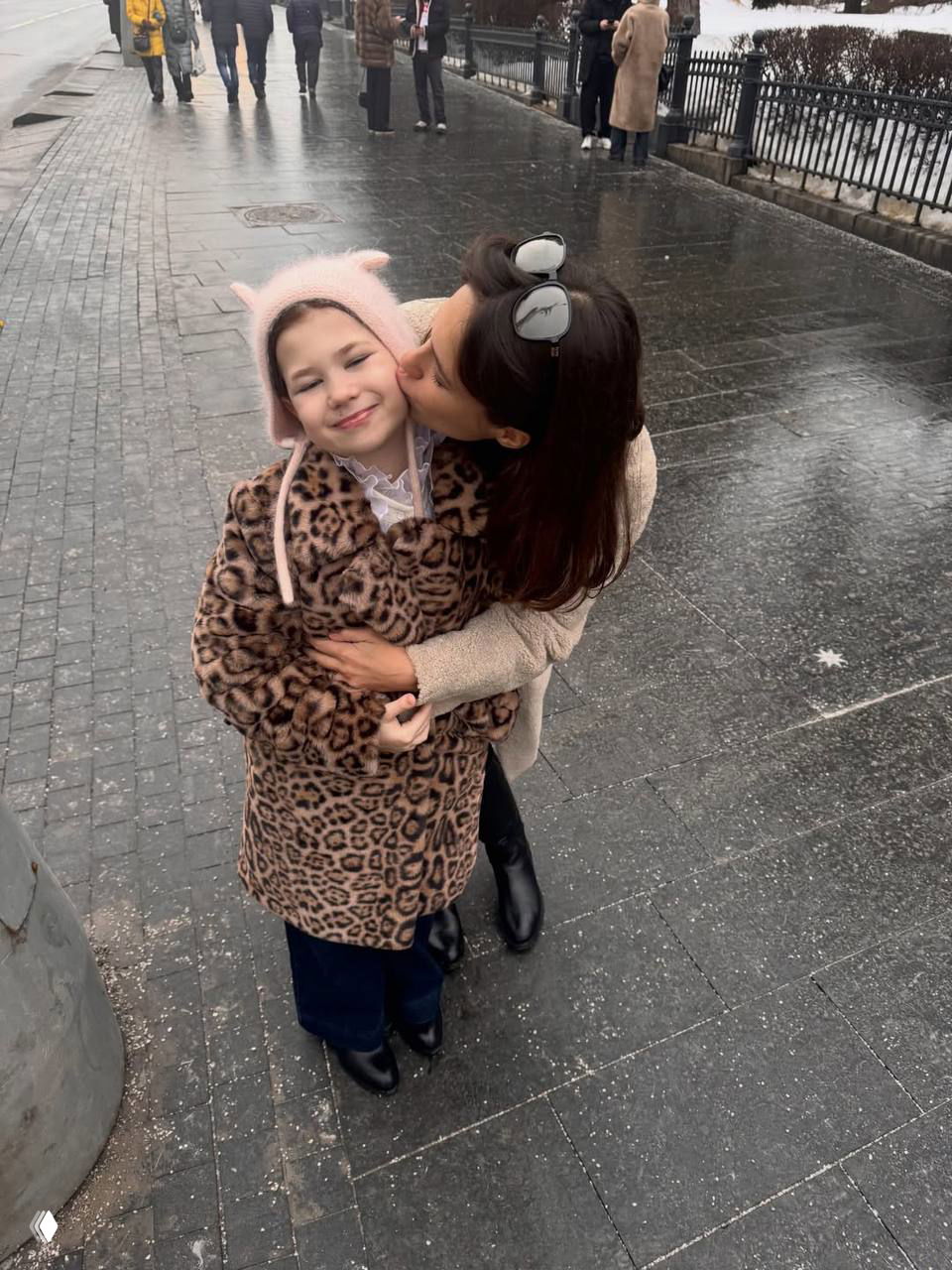 Уличный кадр: женщина целует пощёчину девочке в леопардовой шубке на тротуаре в городском пейзаже, зрители на заднем плане