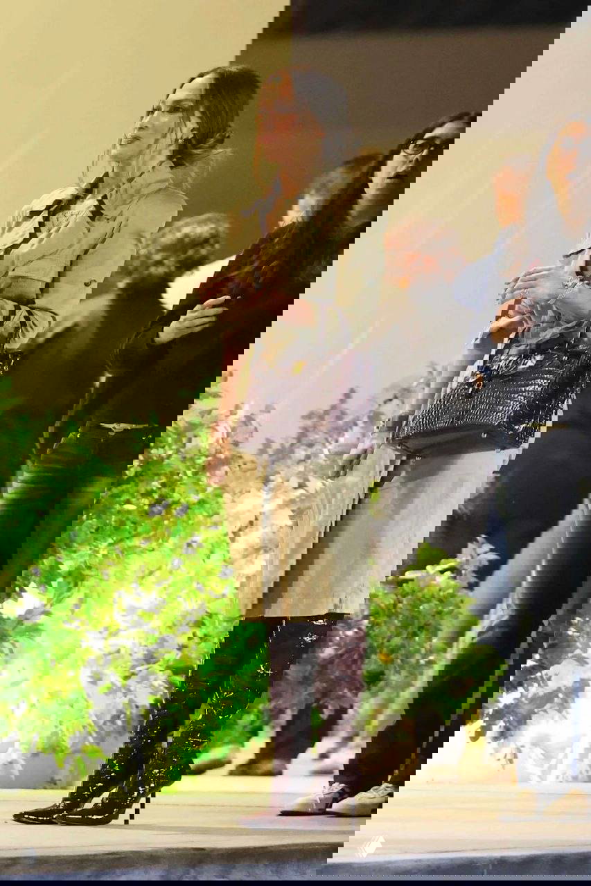 Полный рост Джей Ло на фоне зелёных растений и толпы: тренч ниже колен, лакированные бордовые сапоги и центральная поза возле сцены вечером.