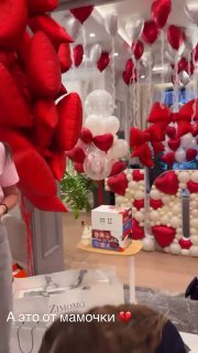 Комната с зеркалами и многочисленными красными шариками‑сердцами, на полу видны пакеты и коробки с подарками на фоне праздничной композиции.