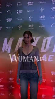 Молодая женщина в повседневном наряде выходит на красную дорожку перед фотоколлом с логотипами и названием картины на заднем плане.