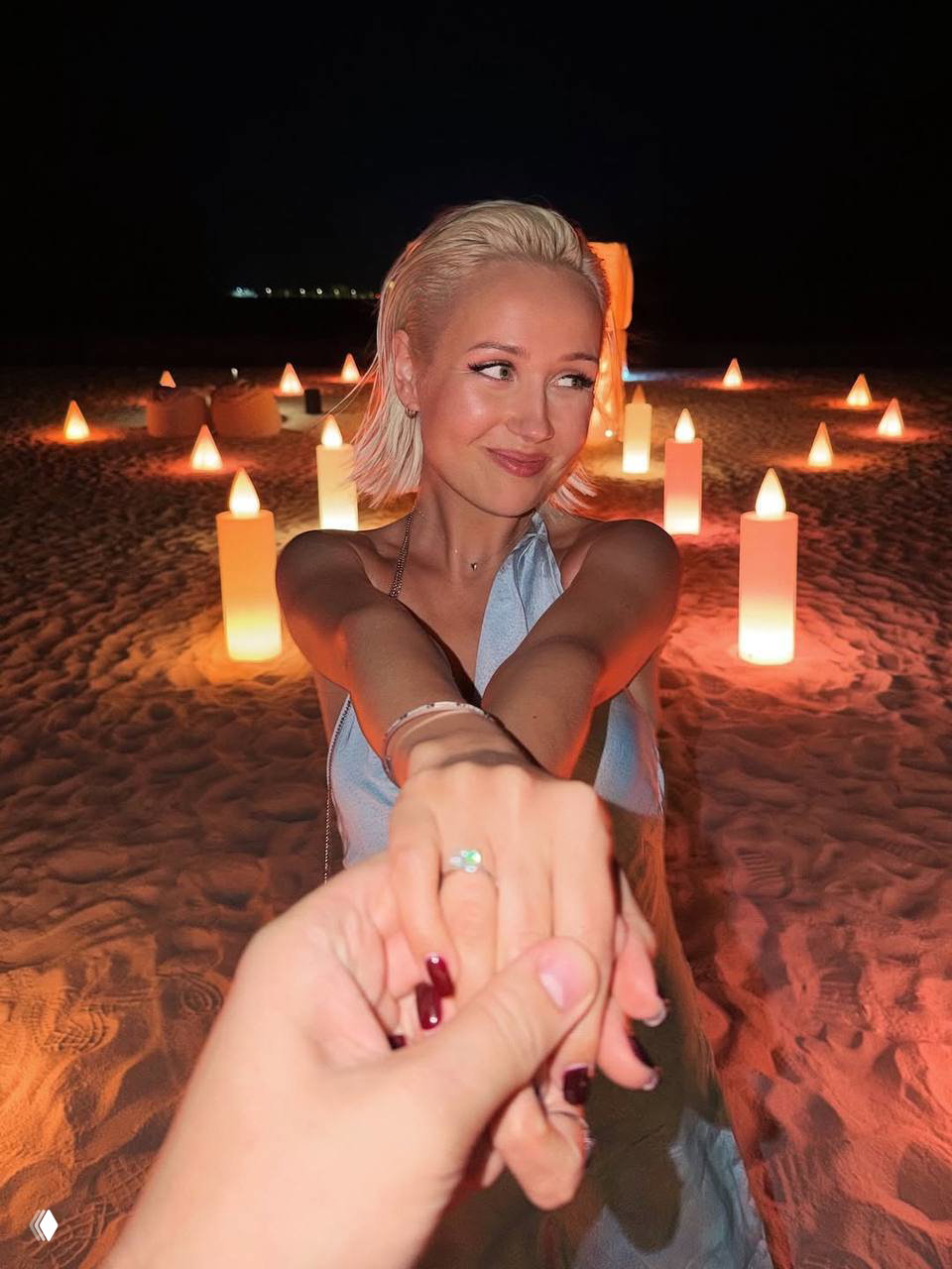 Женщина на ночном пляже с расставленными светящимися свечами тянет руку к камере, демонстрируя помолвочное кольцо.