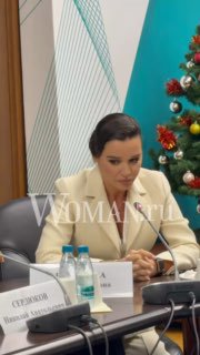 Ксения Бородина в деловом костюме сидит за столом на официальной встрече; на заднем плане новогодняя ёлка и водяной знак Woman.ru, кадр с мероприятия.