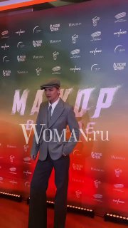 Мужчина в светлом костюме и кепке стоит на фоне бренд-стены с названием фильма, фотозона премьеры выполнена в тёплых тонах.