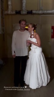Ольга Бузова в свадебном платье стоит рядом с мужчиной на фоне служебного или закулисного пространства — кадр выглядит как со съёмок или мероприятия.