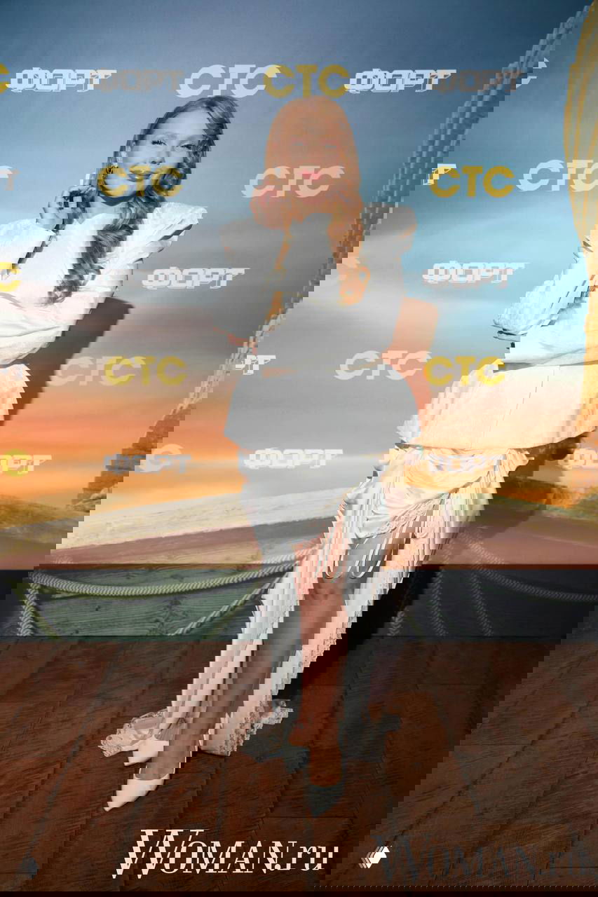 Женщина в белом наряде и туфлях позирует на ковровой дорожке премьеры, фон с фирменными логотипами шоу и телеканала