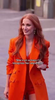 Женщина с длинными рыжими волосами в ярком оранжевом костюме, улыбается и идёт по улице перед камерами — кадр со светского события.