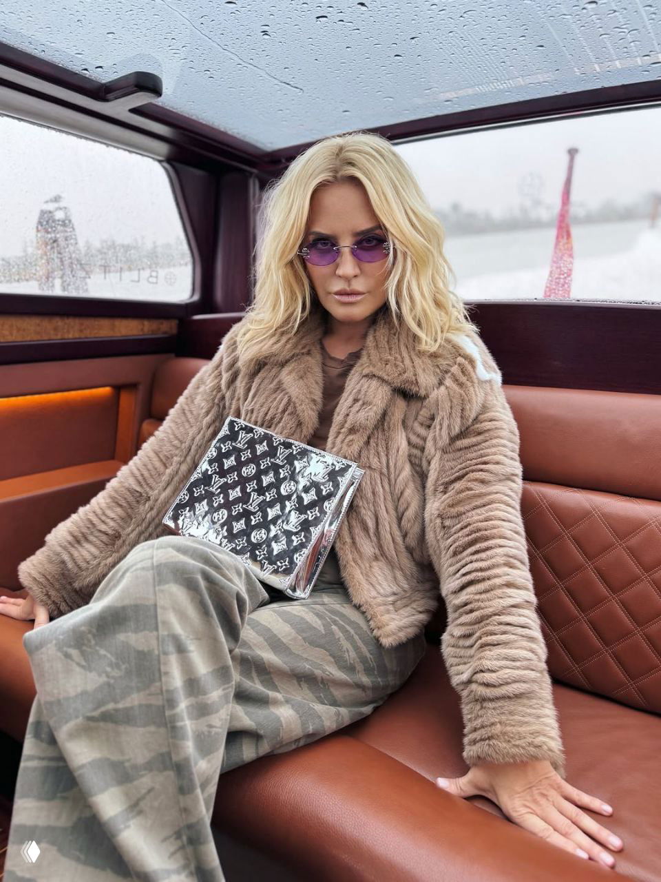 Женщина в плюшевой светло-коричневой шубке и фиолетовых очках сидит в каюте лодки, рядом виднеется дождливая река и городской пейзаж.